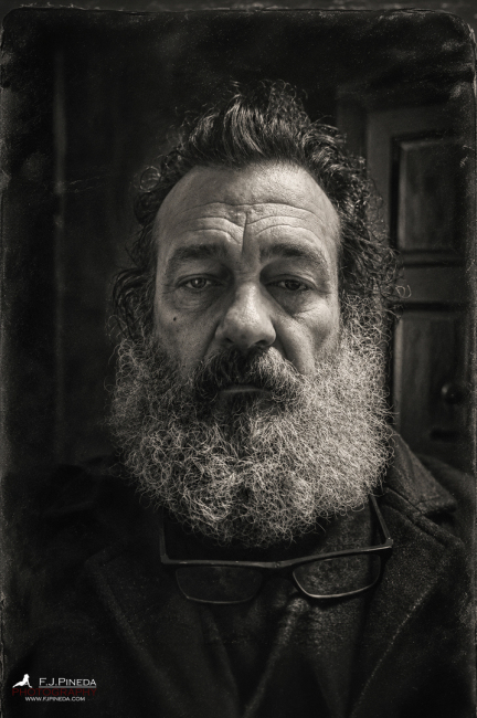 Retrato en blanco y negro de un hombre con barba abundante y gafas al cuello, frente a una puerta cerrada, en una atmósfera oscura e introspectiva.