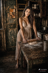Chloe sentada junto a una mesa de madera en una cocina antigua, con vestido claro y luz cálida, serie Las Habitaciones de Chloe de F. J. Pineda.