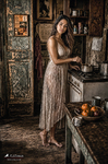 Chloe de pie en una cocina antigua, junto a la encimera y los utensilios, con vestido claro y ambiente cálido.