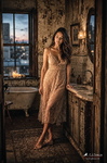 Chloe de pie junto a un lavabo antiguo, con vestido claro y luz cálida, en una escena íntima de Las Habitaciones de Chloe.