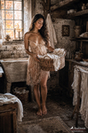 Chloe de pie en una estancia antigua sosteniendo una cesta de ropa, con vestido claro y luz cálida.