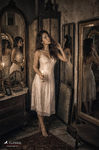 Chloe de pie ante unos espejos o marcos en una habitación antigua, con vestido claro y ambiente íntimo, dentro de la serie Las Habitaciones de Chloe.