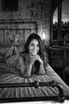 Toma falsa en blanco y negro de Chloe recostada bajo una manta o sábana, mirando a cámara en una habitación antigua.
