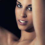 Retrato de mujer adulta con los brazos levantados, mirada directa, piercing en la nariz y fondo oscuro.