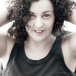 Retrato de mujer adulta con cabello rizado y las manos levantadas hacia la cabeza, sobre fondo claro.
