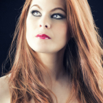 Retrato de mujer adulta pelirroja con vestido rojo, mirada elevada y fondo oscuro.