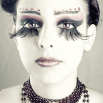 Retrato frontal de mujer adulta con maquillaje artístico, pestañas largas, pedrería sobre las cejas y collar oscuro.