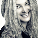Retrato en blanco y negro de mujer adulta rubia sonriendo, con tatuaje visible en el hombro.