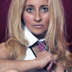Retrato de mujer adulta rubia con camisa blanca, corbata rosa y fondo rojo oscuro.