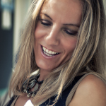 Retrato de mujer adulta rubia sonriendo mientras mira hacia abajo, con luz natural en un interior.