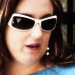Retrato de mujer adulta con gafas de sol blancas, pendientes de perla y jersey azul.