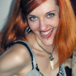 Retrato de mujer adulta pelirroja sonriendo, con piercing en el labio y collar con colgante.