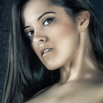 Retrato de mujer adulta con hombros desnudos y cabello oscuro, mirando a cámara sobre un fondo oscuro y texturizado.