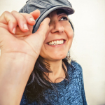 Retrato de mujer adulta sonriendo con gorra azul y jersey del mismo tono, levantando la mano hacia la visera.