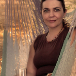 Retrato de mujer adulta sentada en una hamaca, con un vaso en la mano y luz cálida de exterior.