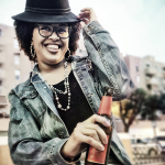 Retrato de mujer adulta con sombrero negro, gafas, chaqueta vaquera y una botella en la mano.