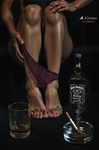 Piernas de una mujer adulta junto a una botella de whisky, un vaso y un cenicero, con una prenda íntima roja entre los pies.