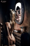 Mujer desnuda sentada frente a un tocador, mirando su reflejo en un espejo ovalado dentro de una habitación tenue.