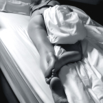 Mujer adulta recostada entre sábanas blancas en una imagen en blanco y negro de luz suave.