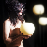 Mujer adulta sosteniendo una esfera o lámpara luminosa entre las manos, sobre fondo oscuro.