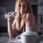 Mujer adulta rubia con lencería negra, sentada a una mesa con una taza y un cigarrillo en la mano.