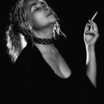 Mujer adulta en blanco y negro, fumando y llevando una gargantilla oscura sobre fondo negro.