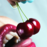 Primer plano de labios femeninos sosteniendo dos cerezas rojas en una imagen de glamour pop.