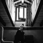 Figura sentada al fondo de una escalera o pasillo en blanco y negro, en una escena de interior más melancólica que sensual.