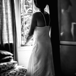 Mujer adulta de espaldas o perfil, vestida con una tela clara, junto a una ventana iluminada en una imagen en blanco y negro.