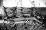 Mujer adulta desnuda tumbada sobre un sofá estampado bajo una ventana iluminada, en una escena de blanco y negro.