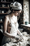 Mujer adulta con delantal y gorro inclinada sobre una mesa enharinada, trabajando la masa en una panadería rústica.