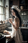 Mujer adulta con delantal claro junto a la encimera, sosteniendo una taza en una escena doméstica de panadería.