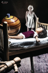 Pequeña figura humana de pie junto a una cama donde yace otra figura, rodeadas por objetos domésticos en miniatura.