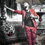 Hombre vestido con chaqueta roja y pantalón de rayas posando junto al agua, con una pequeña nariz roja suspendida cerca.