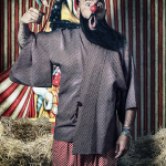 Hombre sujetando un traje gris sin cuerpo visible, en un escenario de circo con fondo de rayas.