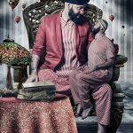 Hombre con chaqueta roja sentado junto a una mesa con objetos antiguos, acompañado por otra figura vestida de rayas.
