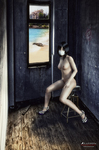 Mujer adulta desnuda junto a una puerta o ventana estrecha, en una escena de interior sombrío vinculada al aislamiento.