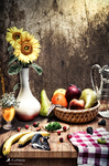 Bodegón luminoso con un girasol en jarrón, cesta de frutas y verduras sobre una mesa cubierta con mantel de cuadros.