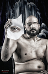 Hombre adulto desnudo o semidesnudo, sentado, sosteniendo una máscara blanca frente al rostro en una escena oscura.