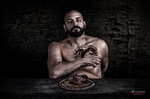 Hombre adulto desnudo con las manos cruzadas sobre el pecho, retratado sobre fondo oscuro con una mesa delante.