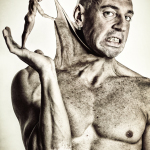 Hombre adulto semidesnudo con expresión tensa, levantando una mano en una pose teatral sobre fondo oscuro.