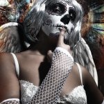 Mujer adulta con maquillaje de calavera, peluca blanca, lencería clara y alas oscuras, llevándose un dedo a los labios en una escena de glamour sombrío.