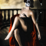 Mujer adulta con maquillaje de calavera, sentada con las piernas cruzadas y una tela roja junto a un sillón oscuro.