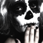 Primer plano de mujer adulta con maquillaje de calavera y cabello claro, tocándose los labios o el rostro con los dedos.