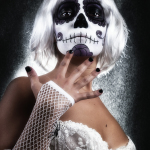 Mujer adulta con maquillaje de calavera, peluca blanca y guante o manga de rejilla, tocándose el cuello o el pecho.