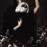 Mujer adulta con maquillaje de calavera y vestido negro, alzando un brazo en una escena teatral con público o figuras al fondo.