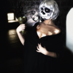 Mujer adulta con maquillaje de calavera y vestido negro, posando de pie en un interior oscuro.