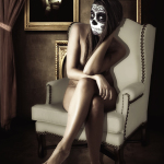 Mujer adulta con maquillaje de calavera sentada en un sillón claro, con las piernas cruzadas y pose elegante.