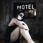 Mujer adulta con maquillaje de calavera sentada en el suelo, abrazándose las piernas, bajo un letrero de motel en una escena sombría.