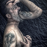 Hombre adulto de perfil, con el brazo tatuado elevado hacia la boca y una mano apoyada en el pecho, en una imagen de tensión corporal.